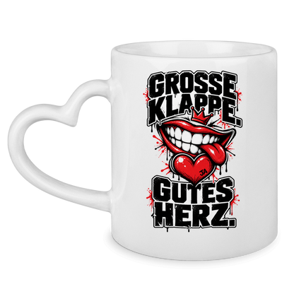 JA Große Klappe, gutes Herz – Tasse mit Herzhenkel in Weiß, Vorderansicht, Aufdruck: Markenspruch „Große Klappe. Gutes Herz.“ mit rotem Mund und Herz